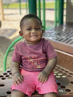 A young child wearing a maroon shirt with words in colorful text and bright pink shorts sits on a playground platform. The background includes playground equipment with green and beige tones.