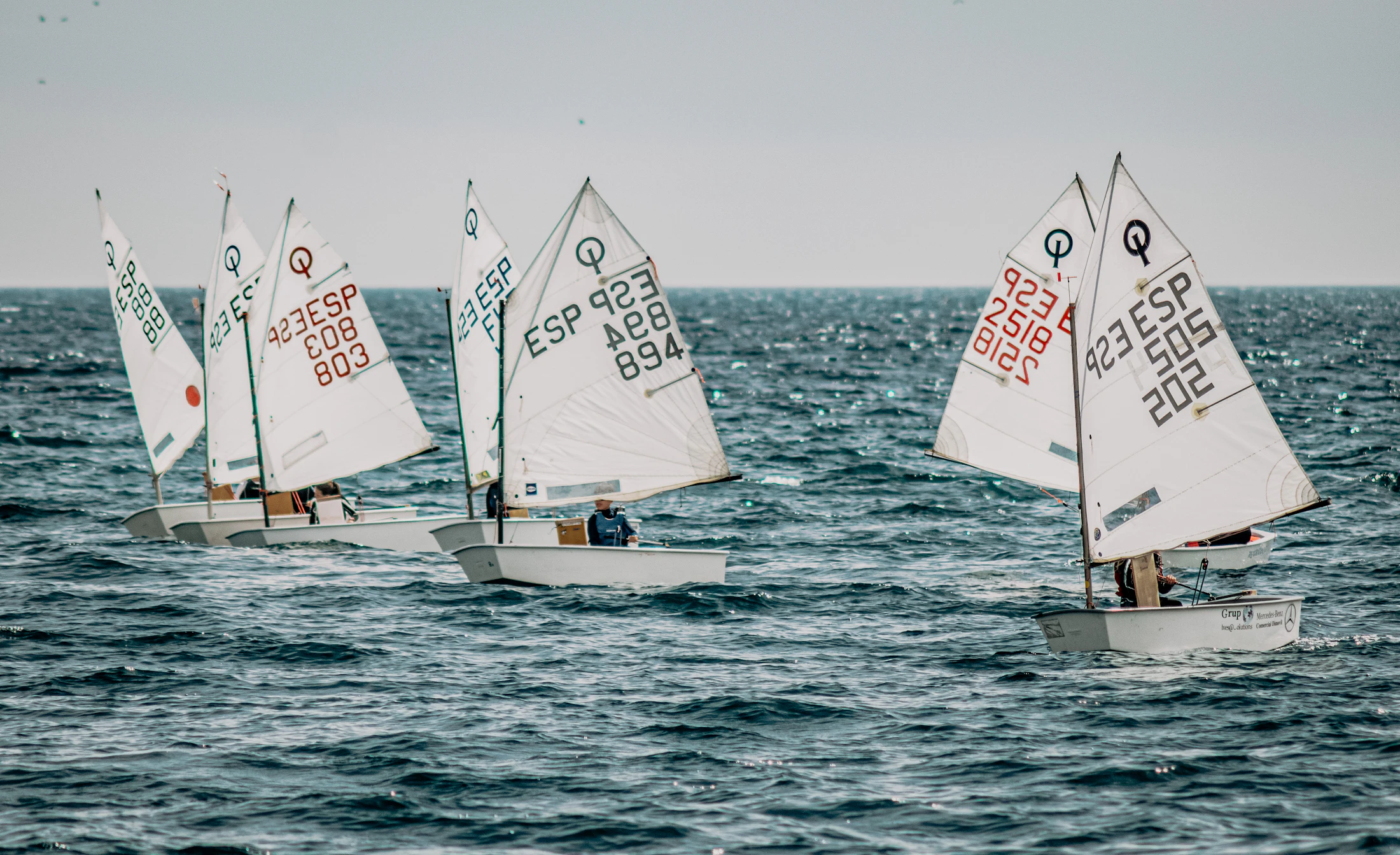 A fleet of small sailing dinghies racing on blue water under a bright sky
