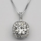 A silver necklace with a round, brilliant-cut diamond pendant surrounded by smaller diamonds arranged in a square halo setting. The pendant is attached to an elegant and delicate silver chain, which complements the sparkle of the diamonds.