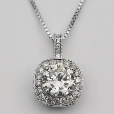 A silver necklace with a round, brilliant-cut diamond pendant surrounded by smaller diamonds arranged in a square halo setting. The pendant is attached to an elegant and delicate silver chain, which complements the sparkle of the diamonds.