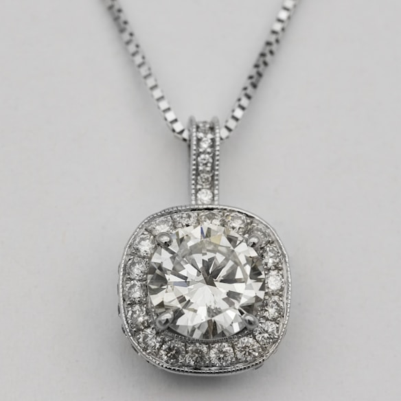 A silver necklace with a round, brilliant-cut diamond pendant surrounded by smaller diamonds arranged in a square halo setting. The pendant is attached to an elegant and delicate silver chain, which complements the sparkle of the diamonds.