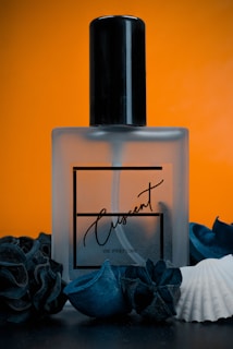 A frosted glass perfume bottle labeled 'Crescent' is centered against a vibrant orange background. The bottle is surrounded by dark blue decorative elements and a white seashell, creating a contrast between the dark and light colors.