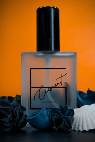 A frosted glass perfume bottle labeled 'Crescent' is centered against a vibrant orange background. The bottle is surrounded by dark blue decorative elements and a white seashell, creating a contrast between the dark and light colors.