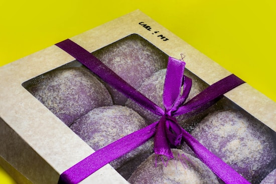 A box containing six round pastries covered with a light dusting of flour is decorated with a vibrant purple ribbon tied into a bow. The box has a transparent lid, allowing the pastries to be visible, and is laid on a bright yellow background.