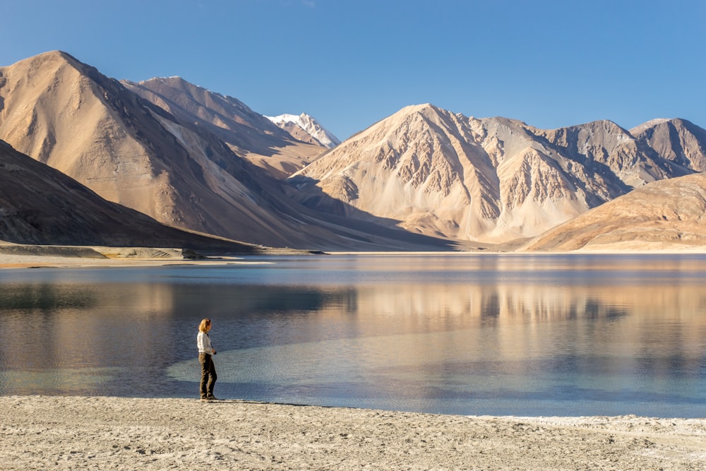 person standing on gray sand near lake and mountains during daytime