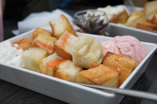 A close-up of a dessert dish featuring toasted bread cubes drizzled with a sweet syrup, accompanied by a scoop of pink ice cream and a dollop of whipped cream. The dish is served in a white rectangular tray.