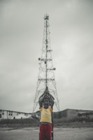 A group of children stand in a line in front of a tall metal tower, with their hands raised in prayer or a symbolic gesture. The background shows overcast skies and a few buildings.