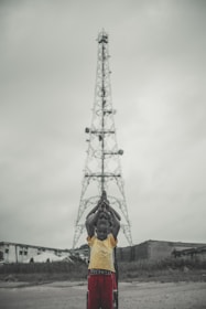 A group of children stand in a line in front of a tall metal tower, with their hands raised in prayer or a symbolic gesture. The background shows overcast skies and a few buildings.