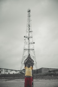 A group of children stand in a line in front of a tall metal tower, with their hands raised in prayer or a symbolic gesture. The background shows overcast skies and a few buildings.