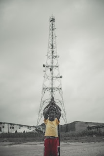 A group of children stand in a line in front of a tall metal tower, with their hands raised in prayer or a symbolic gesture. The background shows overcast skies and a few buildings.