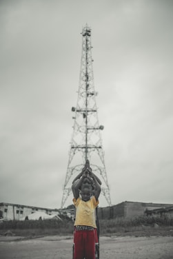 A group of children stand in a line in front of a tall metal tower, with their hands raised in prayer or a symbolic gesture. The background shows overcast skies and a few buildings.