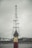 A group of children stand in a line in front of a tall metal tower, with their hands raised in prayer or a symbolic gesture. The background shows overcast skies and a few buildings.