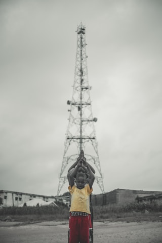 A group of children stand in a line in front of a tall metal tower, with their hands raised in prayer or a symbolic gesture. The background shows overcast skies and a few buildings.