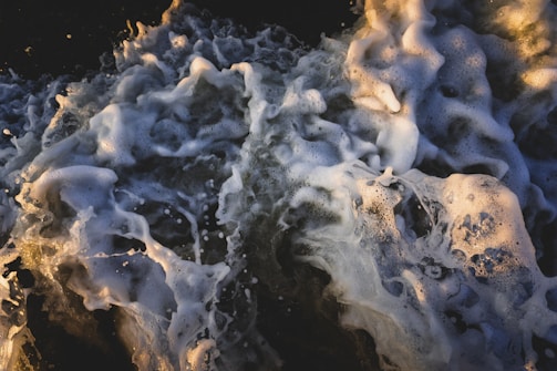 Foamy ocean waves crash and churn with a mix of light and dark water. The frothy white foam creates intricate patterns against the deep blue and green shades of the sea. The light highlights certain areas of the foam, giving depth and texture to the scene.