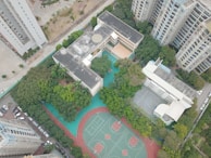 An aerial view of a complex featuring a large building surrounded by greenery, with sports facilities including basketball courts and a running track. Several high-rise apartment buildings are visible nearby, along with parked cars along the streets.
