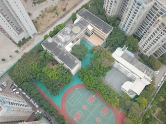 An aerial view of a complex featuring a large building surrounded by greenery, with sports facilities including basketball courts and a running track. Several high-rise apartment buildings are visible nearby, along with parked cars along the streets.