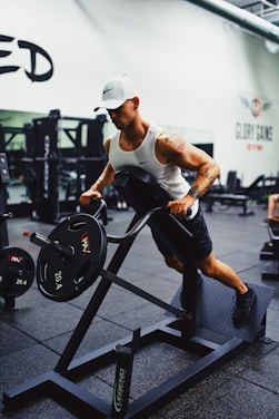 A person is working out in a gym using a weight machine. They are wearing a white tank top, black shorts, and a white cap. The gym is equipped with various fitness machines, and there are weights visible in the background.