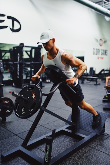 A person is working out in a gym using a weight machine. They are wearing a white tank top, black shorts, and a white cap. The gym is equipped with various fitness machines, and there are weights visible in the background.