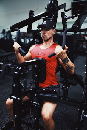 A man is exercising on a chest press machine at a gym. He is wearing a red sleeveless shirt, black shorts, and a black cap. His arms are engaged in pushing the machine handles away from his body. The gym is equipped with various fitness equipment, and the setting is indoors with a focused and determined atmosphere.