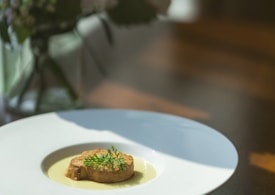 A dish is elegantly presented on a large white plate. The central focus is a piece of food, possibly roasted meat or a vegetable terrine, topped with fresh green herbs. It sits in a small amount of creamy, light-colored sauce. The background is softly blurred with hints of greenery and a soft, warm lighting stream, suggesting a relaxed dining experience.