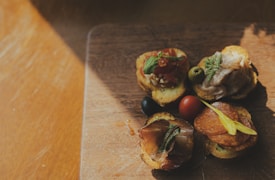 A close-up view of a wooden board topped with various appetizers including slices of bread with toppings such as cured meats, olives, and garnishes. Sunlight casts shadows across the board, adding a warm and rustic feel.