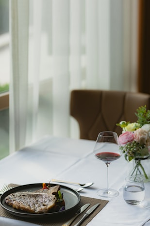 A sophisticated dining setup features a plate with a cooked steak served on a clean white tablecloth. Beside the plate, there's a glass of red wine and a small glass of water. Elegant cutlery is arranged neatly on a brown placemat. In the background, a flower arrangement with pastel blooms adds a touch of elegance to the setting.