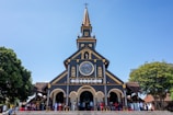 A large wooden church with intricate architecture stands against a clear blue sky. The structure features a tall, pointed steeple adorned with a cross. The facade includes a large circular stained glass window and several arched doorways. People are gathered at the entrance, some sitting on red stools, and trees flank both sides of the church.