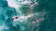 An aerial view captures three surfers paddling through the turquoise ocean. The waves create white foam as they break, contrasting with the deep green-blue of the water. The surfers are scattered, each on their surfboards, seemingly in coordination to catch the next wave.