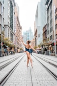 A ballerina stands on pointe in the middle of a city street, surrounded by tall buildings. She is wearing a dark leotard and has her arm gracefully extended upward. The street is lined with shops and there are people in the background.
