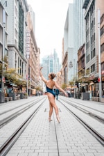 A ballerina stands on pointe in the middle of a city street, surrounded by tall buildings. She is wearing a dark leotard and has her arm gracefully extended upward. The street is lined with shops and there are people in the background.