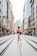 A ballerina stands on pointe in the middle of a city street, surrounded by tall buildings. She is wearing a dark leotard and has her arm gracefully extended upward. The street is lined with shops and there are people in the background.