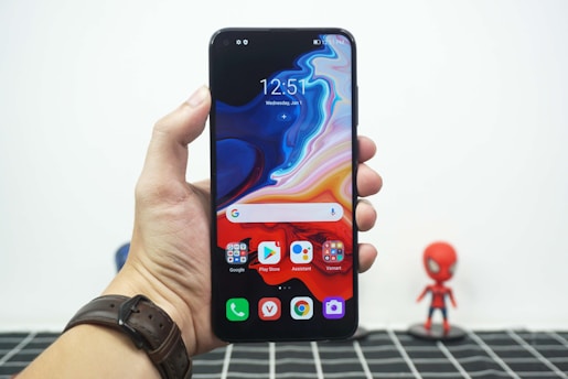 A hand holding a smartphone with a colorful, abstract wallpaper displayed on its screen. The phone interface shows app icons including Google, Play Store, Assistant, and Vsmart. The hand is wearing a brown leather watch. In the blurred background, there is a small figurine resembling a superhero.