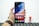 A hand holding a smartphone with a colorful, abstract wallpaper displayed on its screen. The phone interface shows app icons including Google, Play Store, Assistant, and Vsmart. The hand is wearing a brown leather watch. In the blurred background, there is a small figurine resembling a superhero.