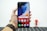 A hand holding a smartphone with a colorful, abstract wallpaper displayed on its screen. The phone interface shows app icons including Google, Play Store, Assistant, and Vsmart. The hand is wearing a brown leather watch. In the blurred background, there is a small figurine resembling a superhero.