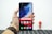 A hand holding a smartphone with a colorful, abstract wallpaper displayed on its screen. The phone interface shows app icons including Google, Play Store, Assistant, and Vsmart. The hand is wearing a brown leather watch. In the blurred background, there is a small figurine resembling a superhero.