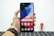 A hand holding a smartphone with a colorful, abstract wallpaper displayed on its screen. The phone interface shows app icons including Google, Play Store, Assistant, and Vsmart. The hand is wearing a brown leather watch. In the blurred background, there is a small figurine resembling a superhero.