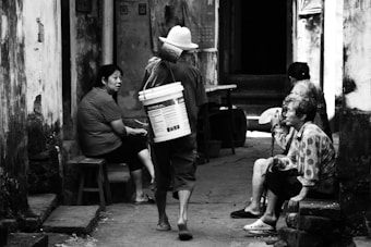 A group of elderly women is sitting and talking in a narrow alley with worn walls. One woman is walking away with a large bucket on her back. The setting has an old, rustic charm.
