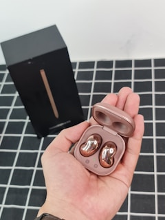 A hand holding a pair of wireless earbuds in a sleek, shiny rose gold color. The earbuds are placed in an open charging case, and the background features a black box and a black-and-white grid-patterned surface.
