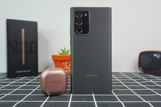 A Samsung smartphone is prominently displayed on a checkered surface with its camera lenses visible. Next to it is a small, round, metallic-looking case, likely for earbuds. In the background, a black box with 'N20' is partially visible, alongside an orange pot with a small green plant and a black object with red accents.