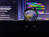 A crystal ball is placed in front of a computer screen displaying colorful code and text. The ball reflects and distorts the vivid lines of code, giving them a swirling, abstract appearance. The background is mostly dark, highlighting the bright neon colors of the text on the screen.