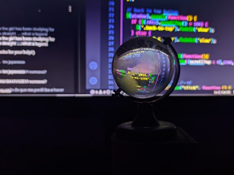 A crystal ball is placed in front of a computer screen displaying colorful code and text. The ball reflects and distorts the vivid lines of code, giving them a swirling, abstract appearance. The background is mostly dark, highlighting the bright neon colors of the text on the screen.