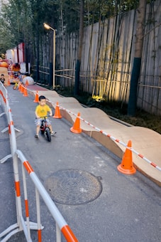 A young child rides a small bike along a pathway lined with orange traffic cones. The path is bordered by a barrier with red and white stripes. Tall bamboo and a lit lamp post create a serene atmosphere along the concrete wall in the background.