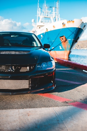 A sleek black car is parked on a pier near a large blue and white ship, under a clear blue sky. The front part of the car is prominently visible, with intricate grill details and shiny components. The ship is anchored close to the shore, with water gently reflecting the sunlight, creating a vibrant scene.