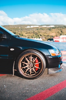 A close-up view of a car, focusing on the front left wheel with bronze-colored alloy rims. The brake calipers are painted red, adding contrast to the wheel. The car is parked on a coastal road with a scenic background of hills, clear sky, and a body of water.