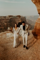 A joyful couple is walking hand-in-hand on a rocky cliff edge, likely celebrating their wedding. The bride is wearing a detailed white gown while the groom is in a light-colored suit. They are both smiling widely, adding to the joyous atmosphere. Behind them, rugged canyon landscapes stretch under a clear sky. An officiant stands in the background near a minimalist ceremony arch decorated with dried flowers.