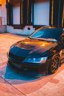 A sleek, black sports car is parked on a concrete surface near a loading dock. The car features a carbon fiber hood and detailed lighting, reflecting ambient sunset tones. Its aerodynamic design and low profile are accentuated by custom wheels.