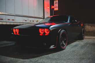 A sleek, matte-black Ford Mustang with a bold front splitter and widebody stance parked in an urban nighttime setting.