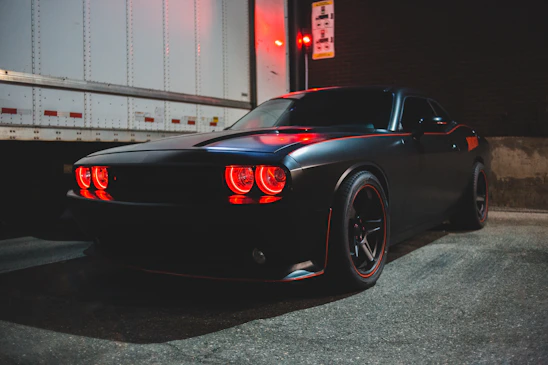 A sleek, matte-black Ford Mustang with a bold front splitter and widebody stance parked in an urban nighttime setting.