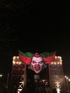 A large, illuminated illustration of a menacing clown face with sharp teeth, red hair, and green accents is mounted at the entrance of a structure, evoking a sense of horror and theatricality. The scene is set at night, with various people visible beneath the entrance, suggesting a festive or event atmosphere.