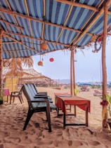 A beachside seating area with wooden chairs and tables covered with red tablecloths is located under a striped blue and white canopy. The sandy beach extends beyond, with other thatched structures and beach loungers scattered in the background.